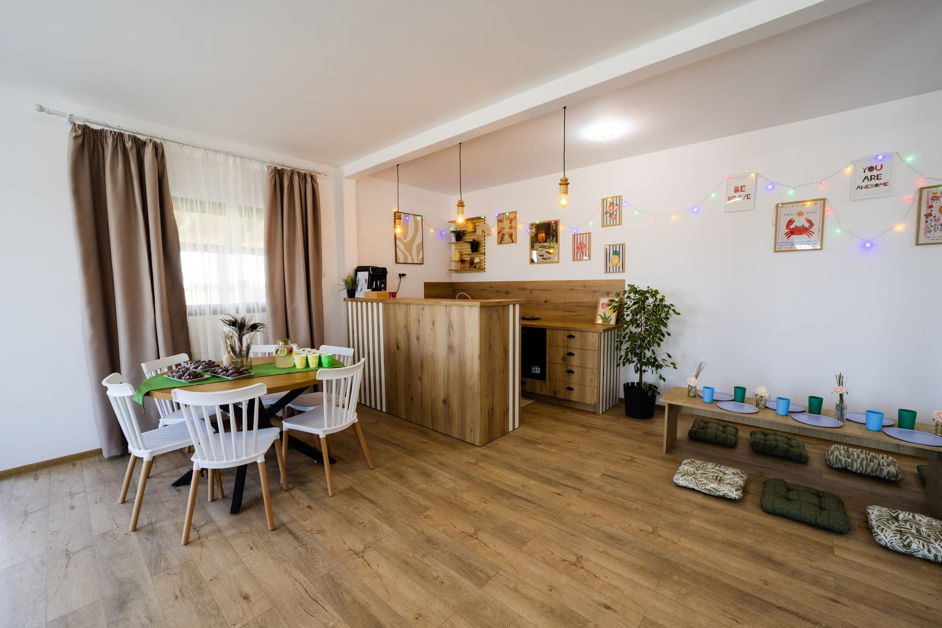 Bar din lemn cu espressor, sertare și frigider, lângă o masă rotundă și masa joasă a copiilor