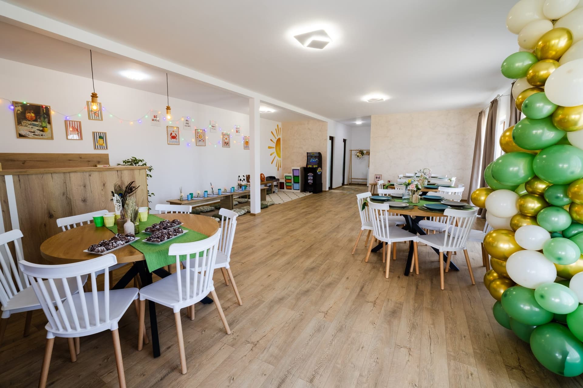 Sala Zi de Poveste pregătită pentru o aniversare: mese rotunde, colț de joacă și baloane verzi cu auriu